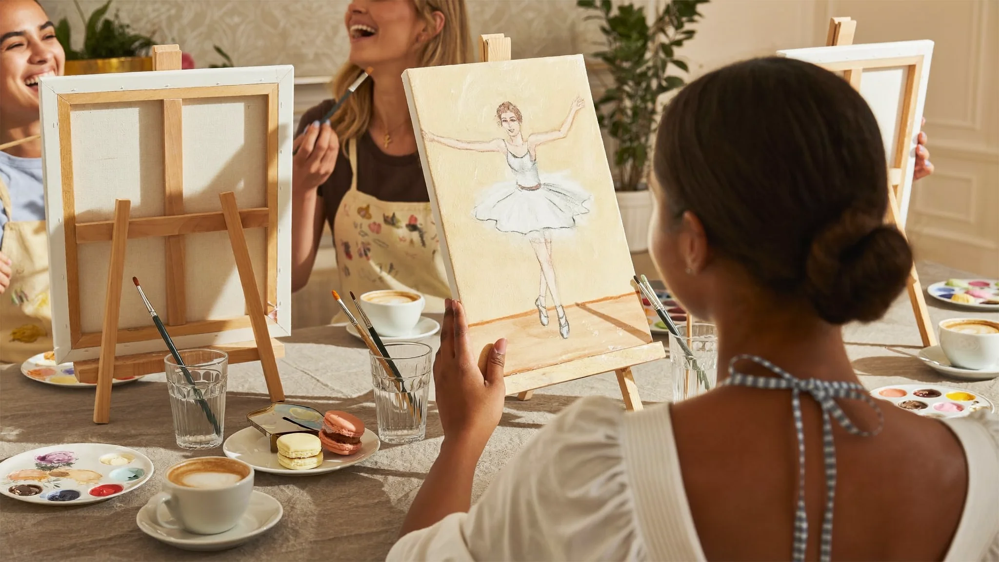 Three friends painting ballet dancer artwork on canvas easels over coffee and macarons at a relaxed morning art session in Dubai